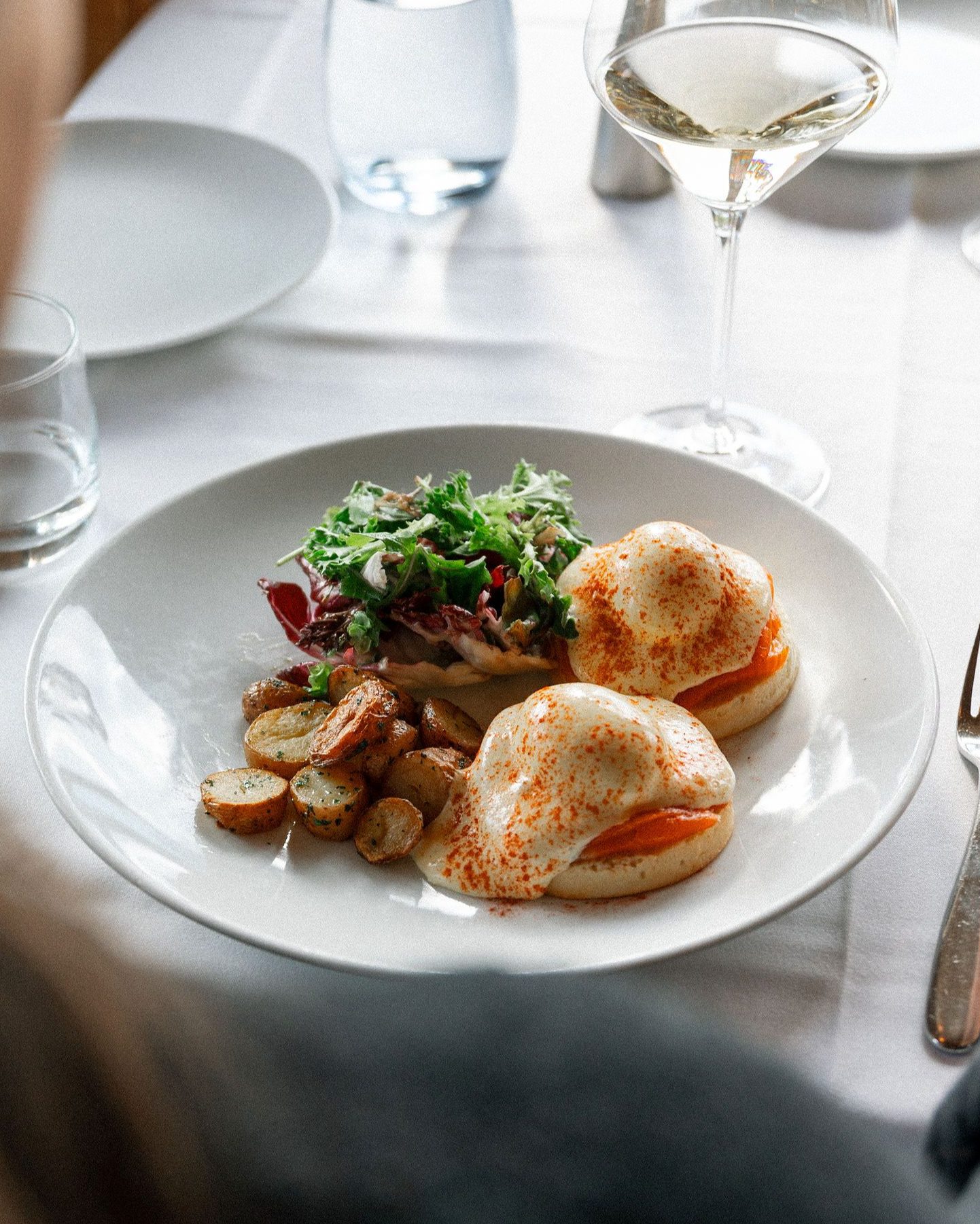 A plate of eggs benedict in front of a glass of white wine.  