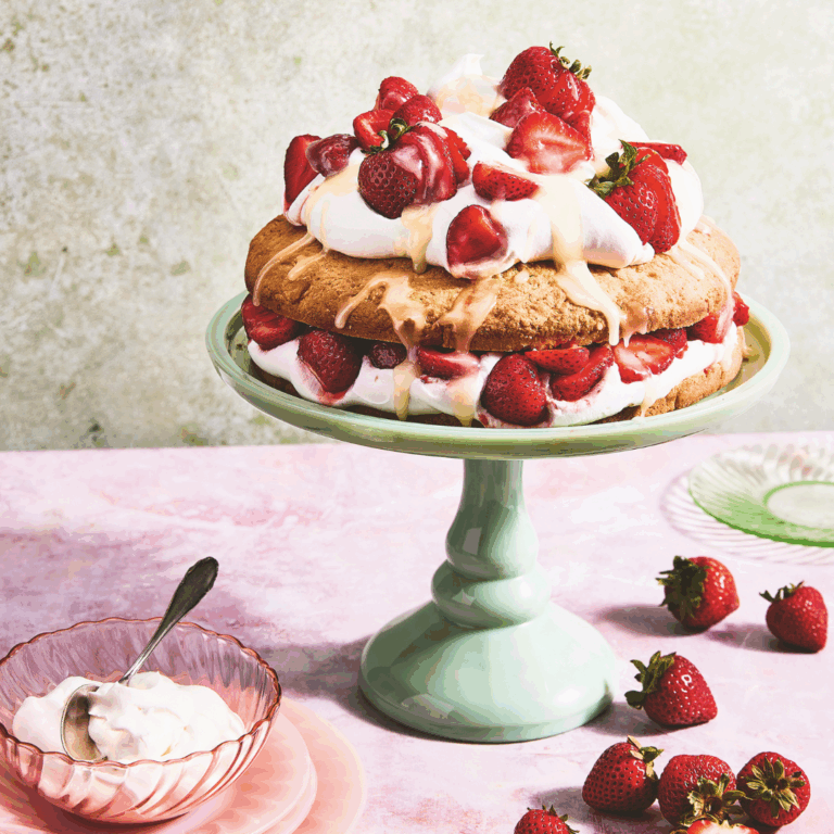 strawberry shortcake layer cake with whipped cream and fresh strawberries ona mint green cake pedestal, surrounded by strawberries and a bowl of whipped cream.