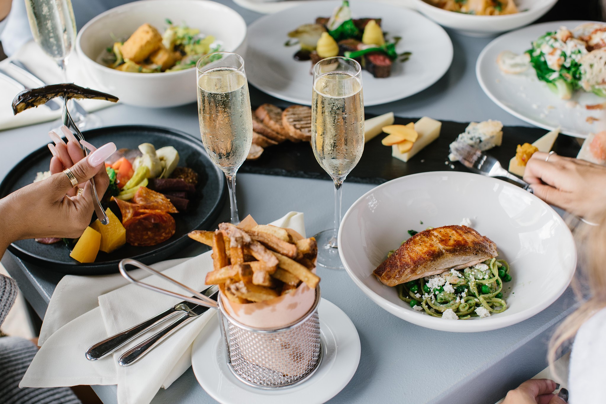 Two flutes of champagne, a cheese board, fries, and other main dishes on a grey table. 