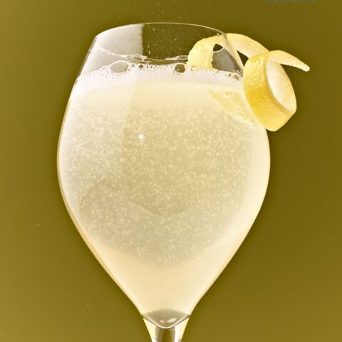 Glass filled with fizzy yellow liquid topped with a lemon twist, in front of an olive green background.