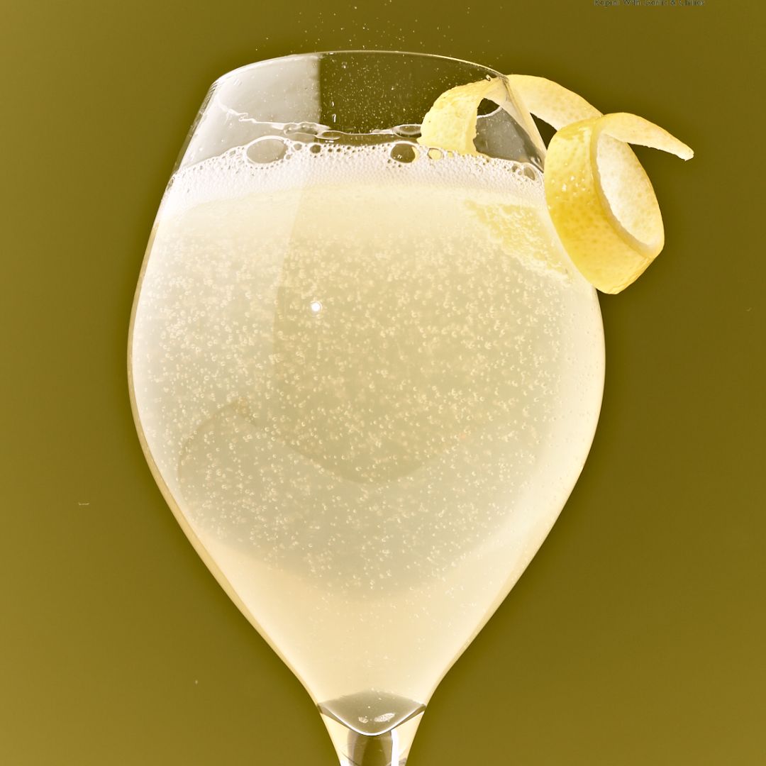 Glass filled with fizzy yellow liquid topped with a lemon twist, in front of an olive green background.