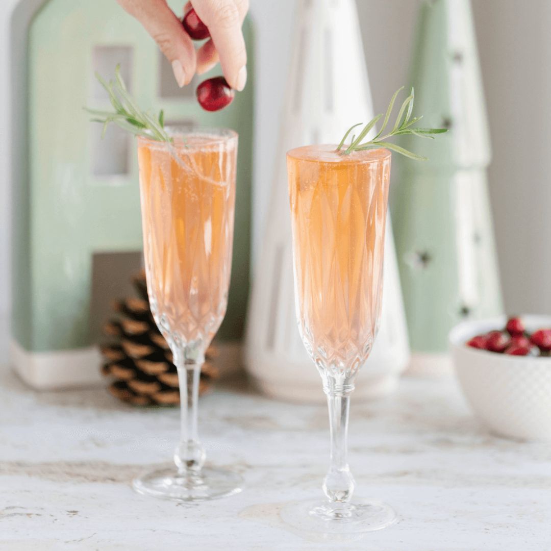 A hand dropping a cranberry into a flute glass filled with light pink liquid. 