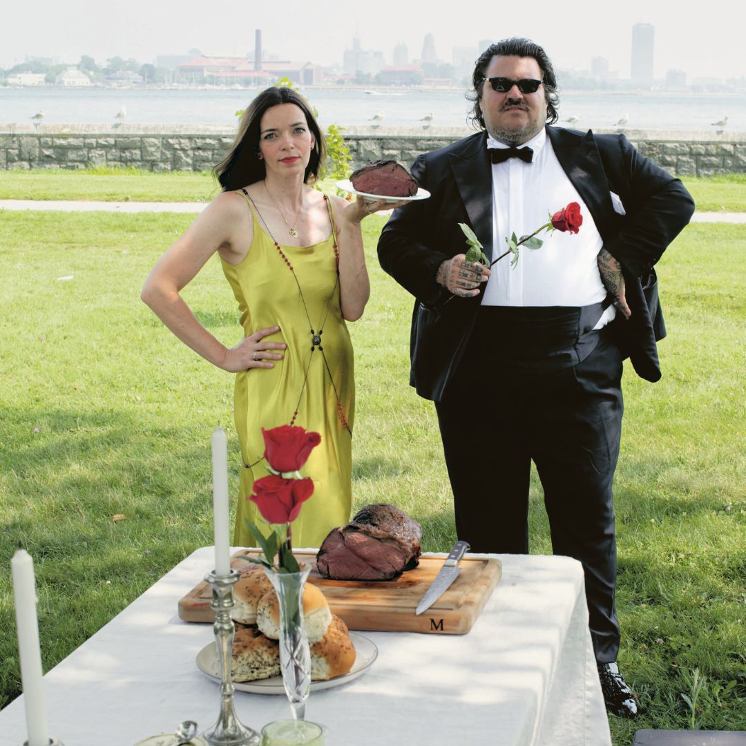 Woman in dress holding a plate next to Matty Matheson wearing a tuxedo while holding a red rose.