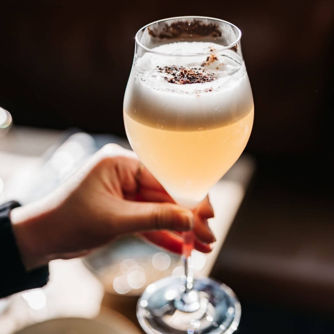 Hand holding a glass filled with light yellow liquid topped with a white foam. 