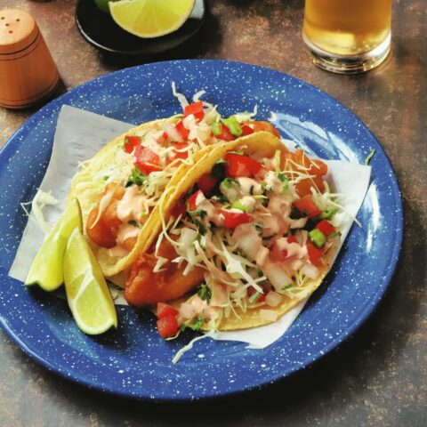 Two baja fish tacos topped with pico de gallo and garnished with two lime wedges.