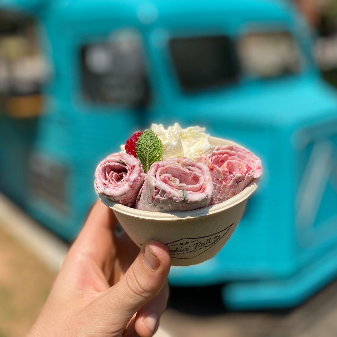 A cup filled with rolled up pink ice cream in front of a blue ice cream truck.