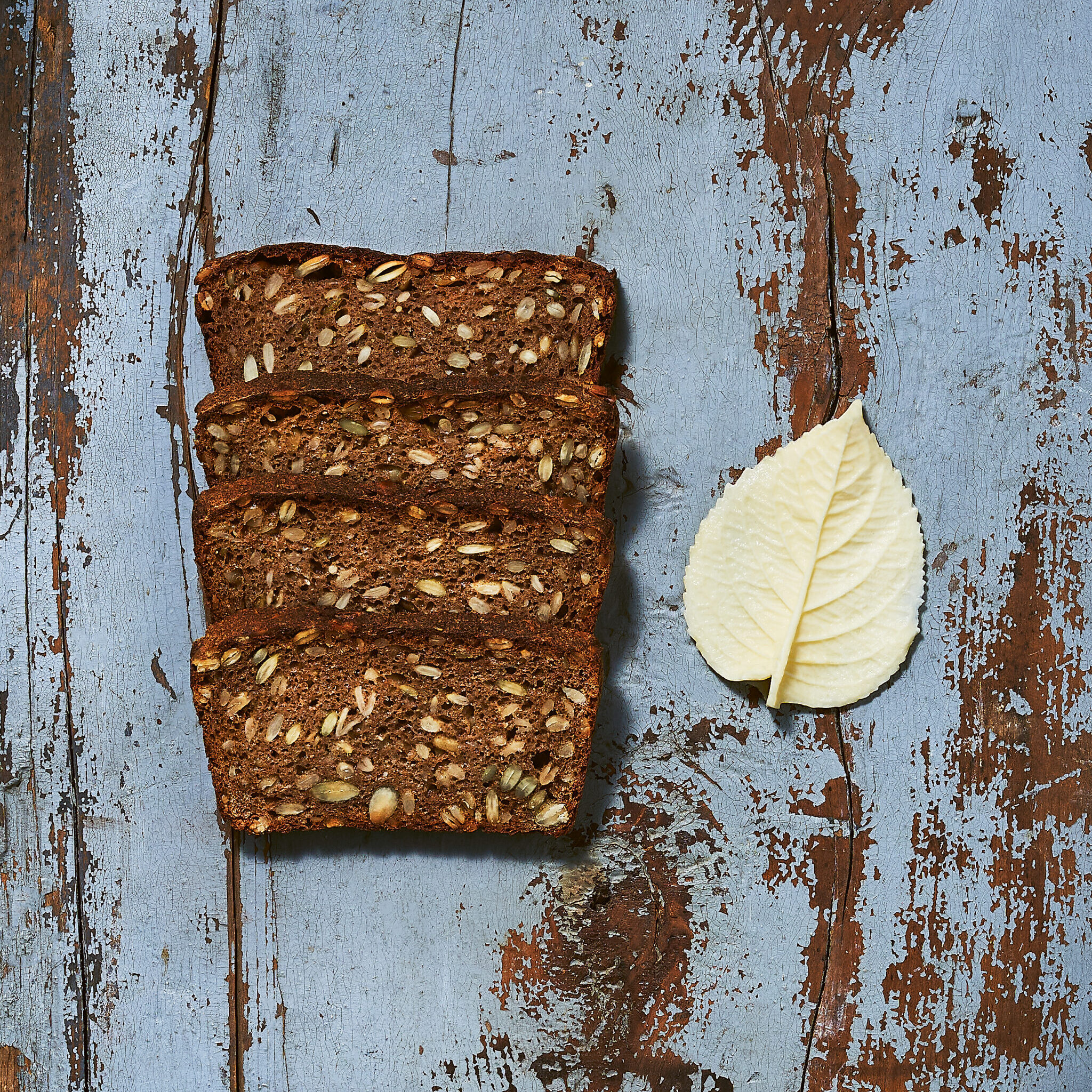 Close-up of four slices of rye bread besides a white leaf on a blue paint-streaked wood background.
