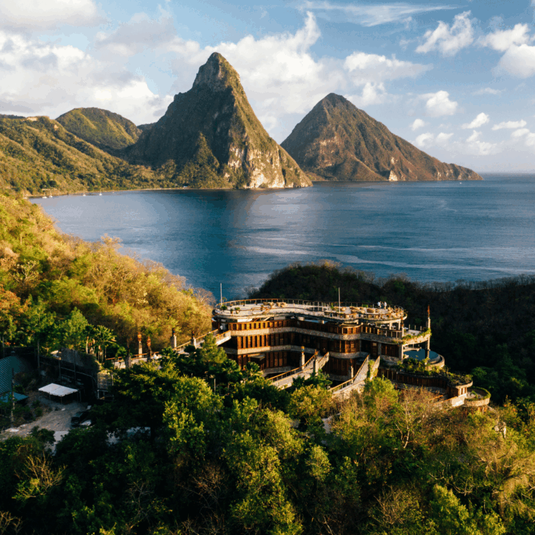 The exterior of a hotel amidst a forest, overlooking water and facing two jagged mountains.