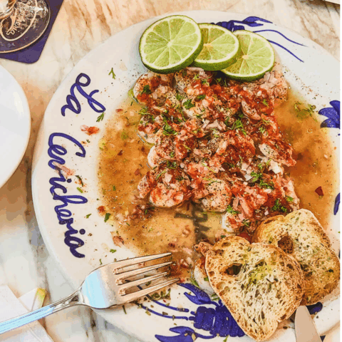 A white plate with "La Cevichería" written on it, holding shrimp ceviche, three slices of lime and two baguette slices.