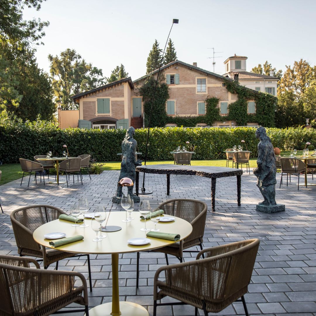 A restaurant's courtyard with tables, chairs and gray statues, with green hedges in the back.