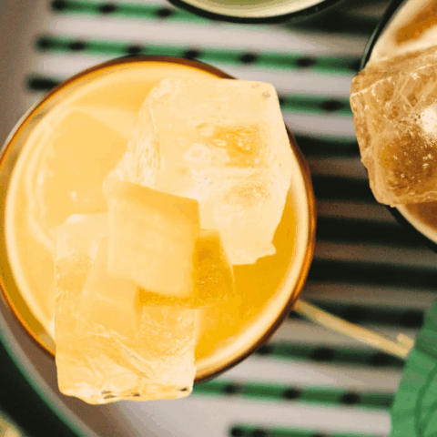 A top angle close-up shot of a glass with a yellow liquid and ice cubes.