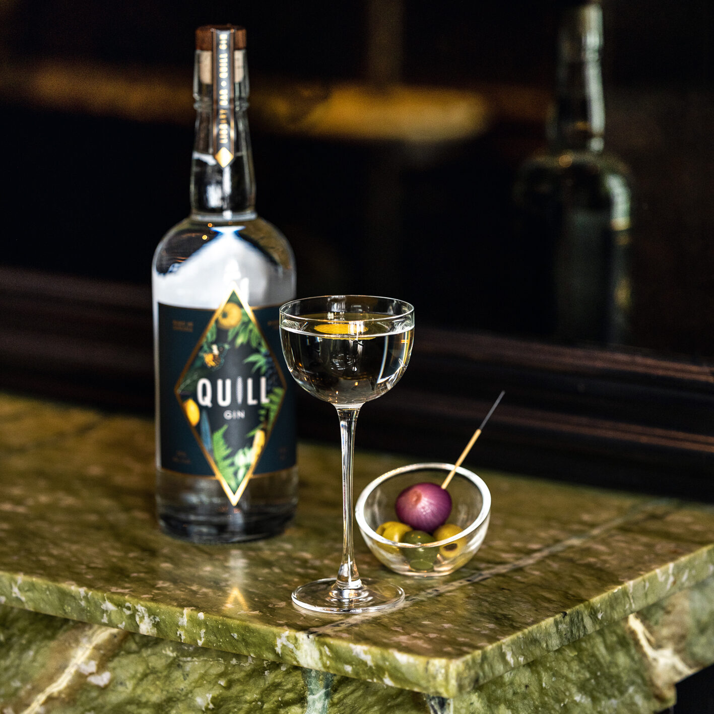 A small bowl of olives, a full pony glass and a bottle of gin on a green marble countertop.