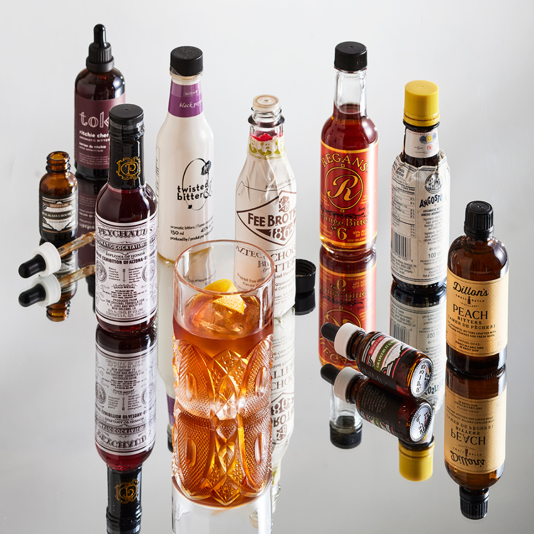 Nine bottles of bitters around a cocktail glass on a mirrored surface.