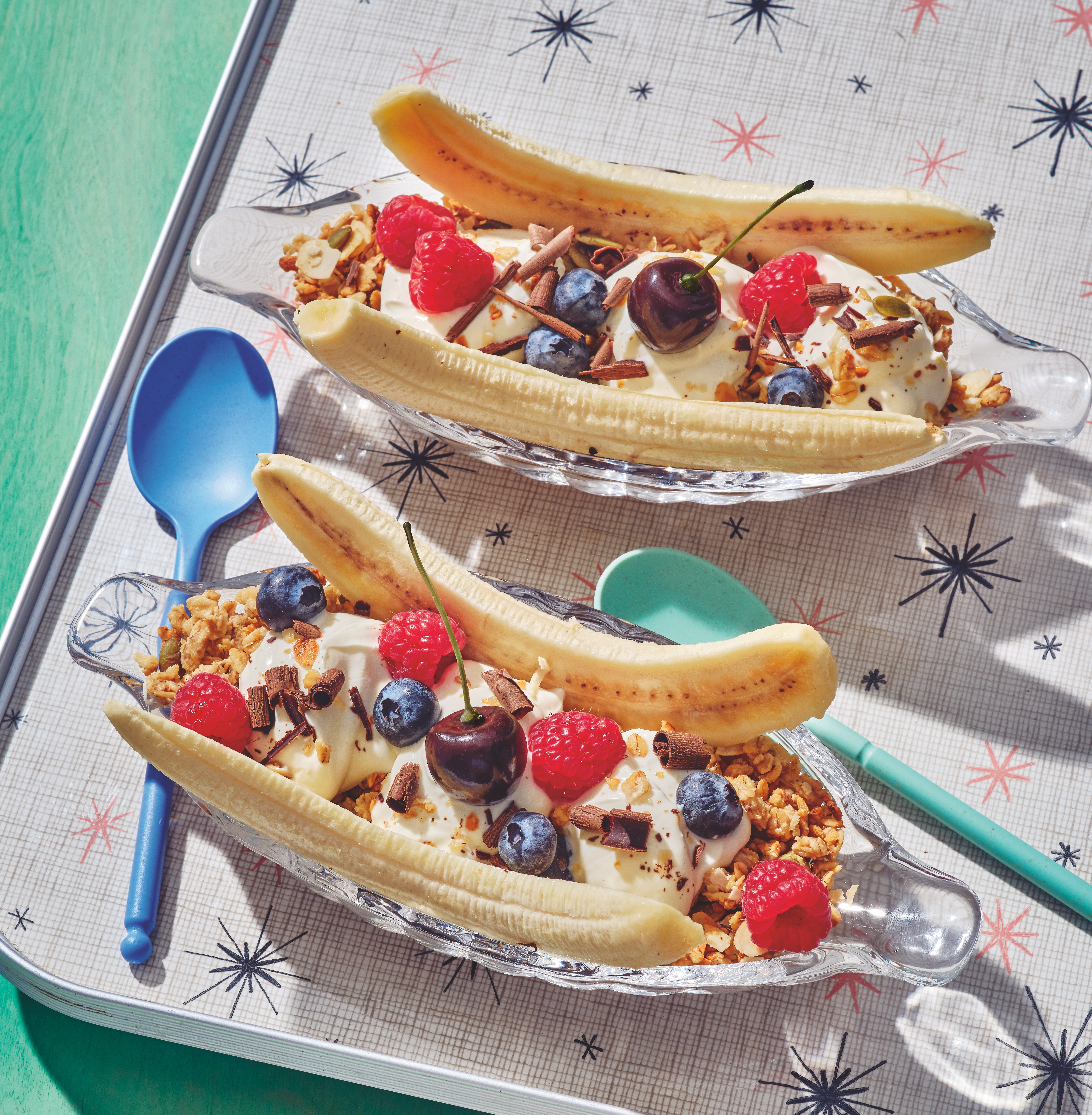 Two banana splits on a bed of granola, topped with yogurt, raspberries, blueberries, chocolate shavings and a red cherry.