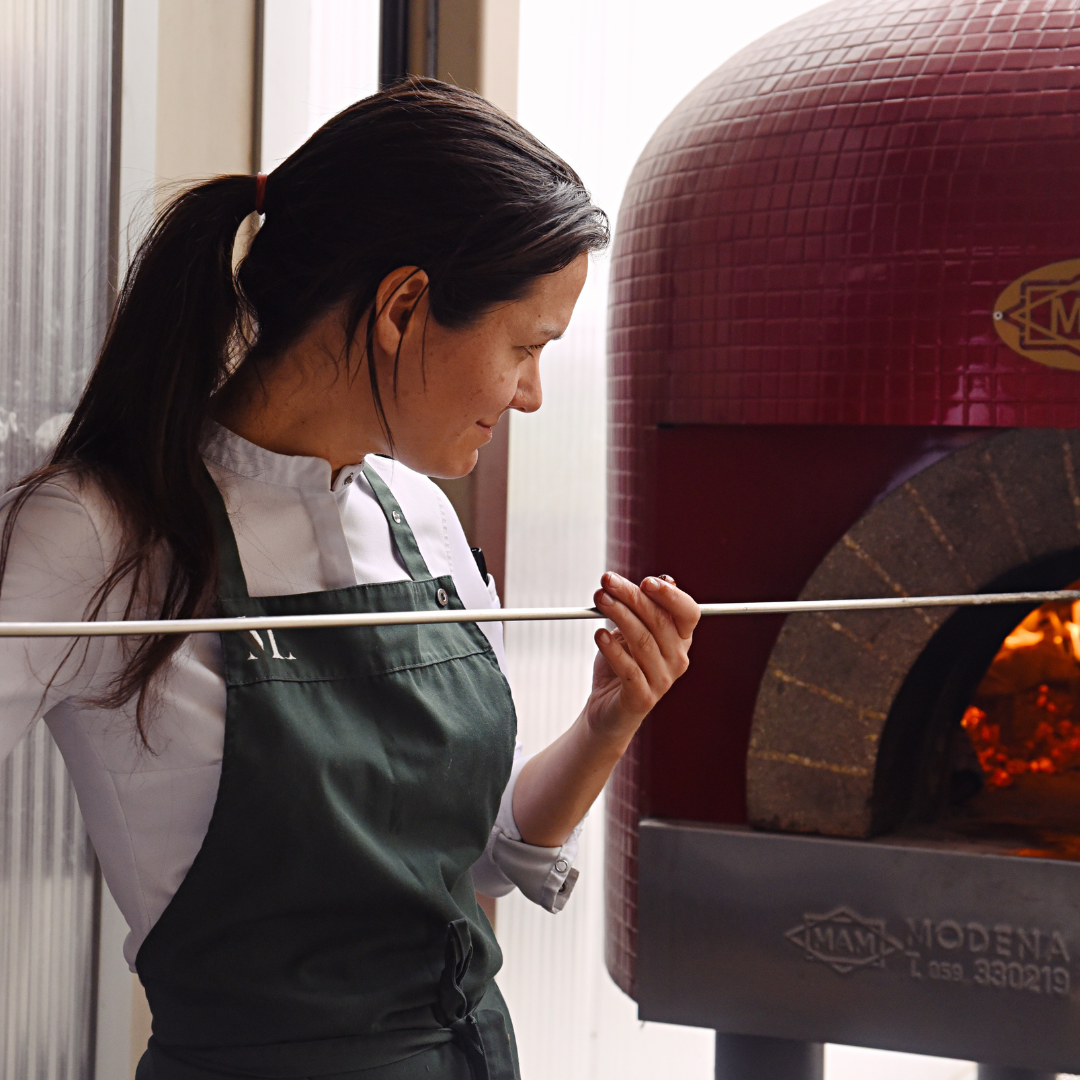 A woman holding a pizza peel going into a wood-fire oven.