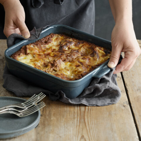 Two hands holding a gray square dish with baked lasagna.