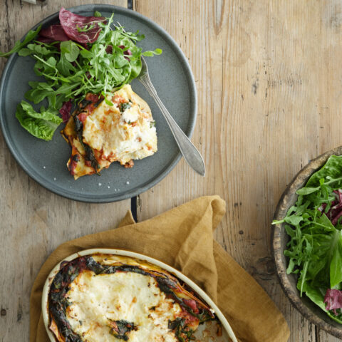 A slice of lasagna and some salad on a gray plate on a wooden background.