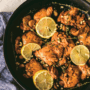 A close-up of a one-pan cooked chicken thighs in a brown sauce, topped with pine nuts and lemon slices.
