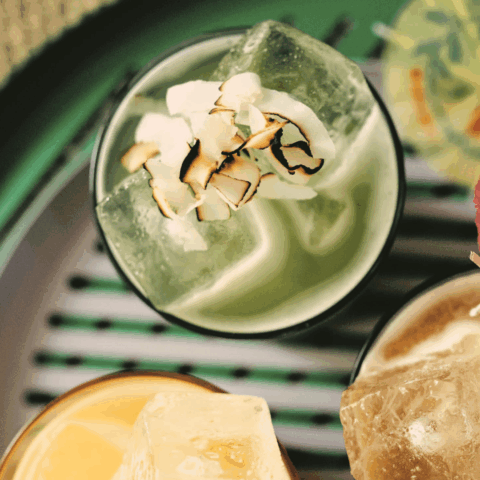 A top angle close-up shot of a glass with a green liquid, ice cubes and toasted coconut flakes.