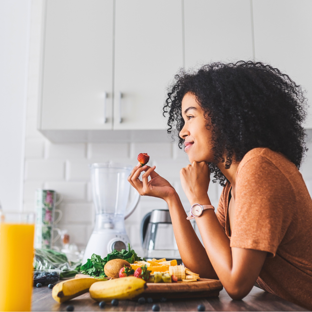 A woman in a modern kitchen leans over a kitchen island and eats a strawberry from a plate of healthy fruit, which includes a colourful variety of fresh produce.