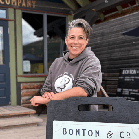 A woman with short gray hair in a gray hoodie in front of an outdoor sign.