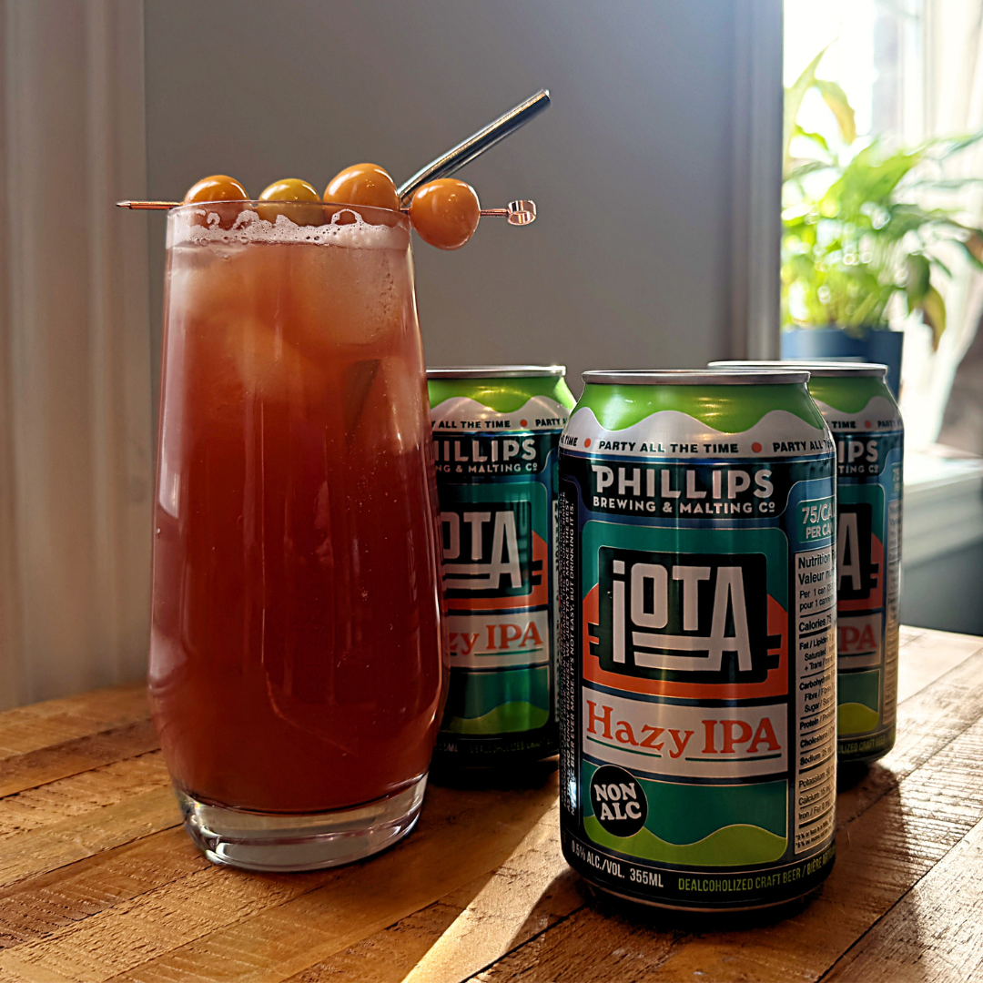 A cocktail glass filled with red drink next to three cans of IPA. 