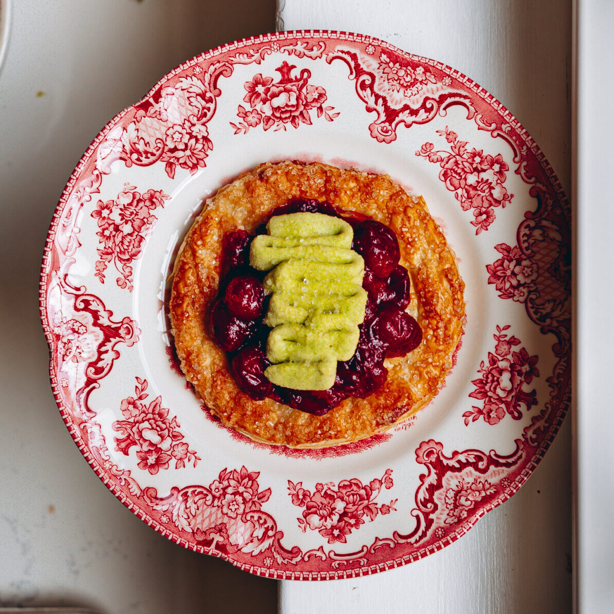 A round piece of baked puff pastry topped with cranberry jam and piped pistachio cream on a red and white decorative plate.