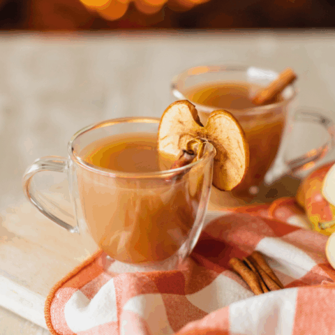 Two mugs of apple cider garnished with cinnamon sticks and dried apple on a marble surface with a fire in the hearth in the background