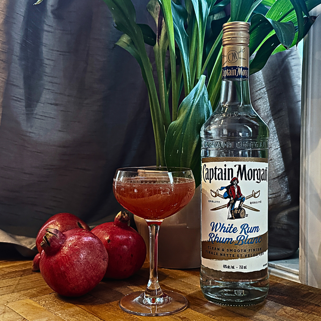 Three pomegranates beside a cocktail in a glass and a clear bottle of white rum. 