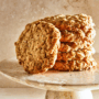 Six chewy oatmeal cookies stacked on top of a marble cake stand on a marble countertop.
