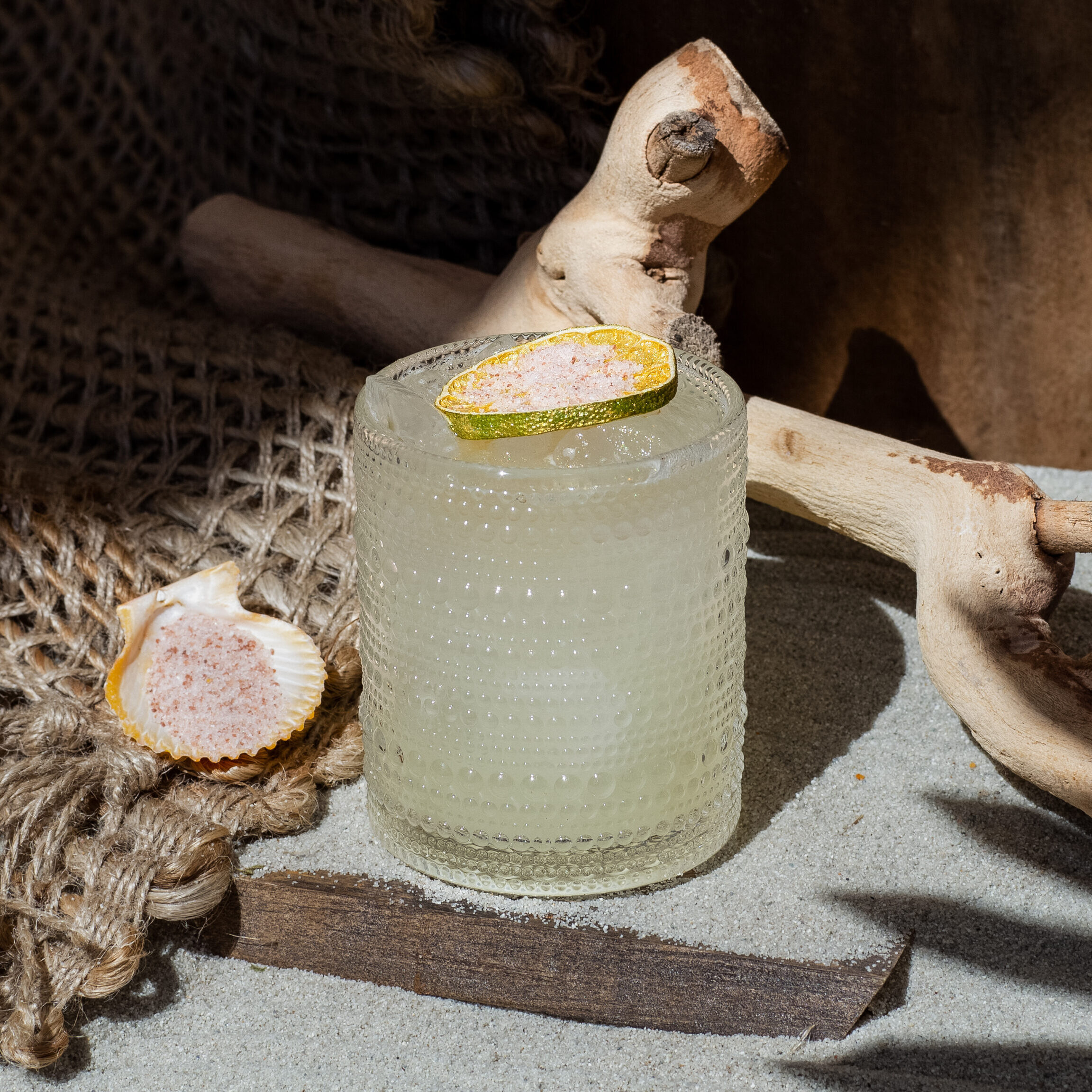 A margarita topped with a lime wheel on a sandy surface with a seashell.
