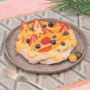 Pavlova topped with an orange sauce, strawberries, blueberries, peach slices next to sliced calamansi on a plate.