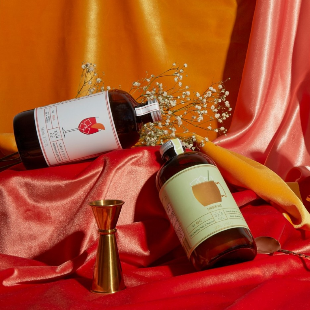 Two bottles of cocktail syrup and a gold jigger on a pink and orange fabric background.