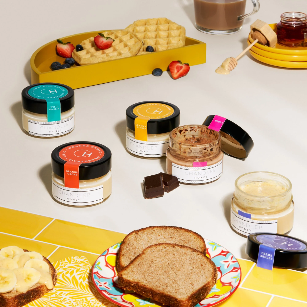 Five jars of honey surrounded by plates of toast and waffles.