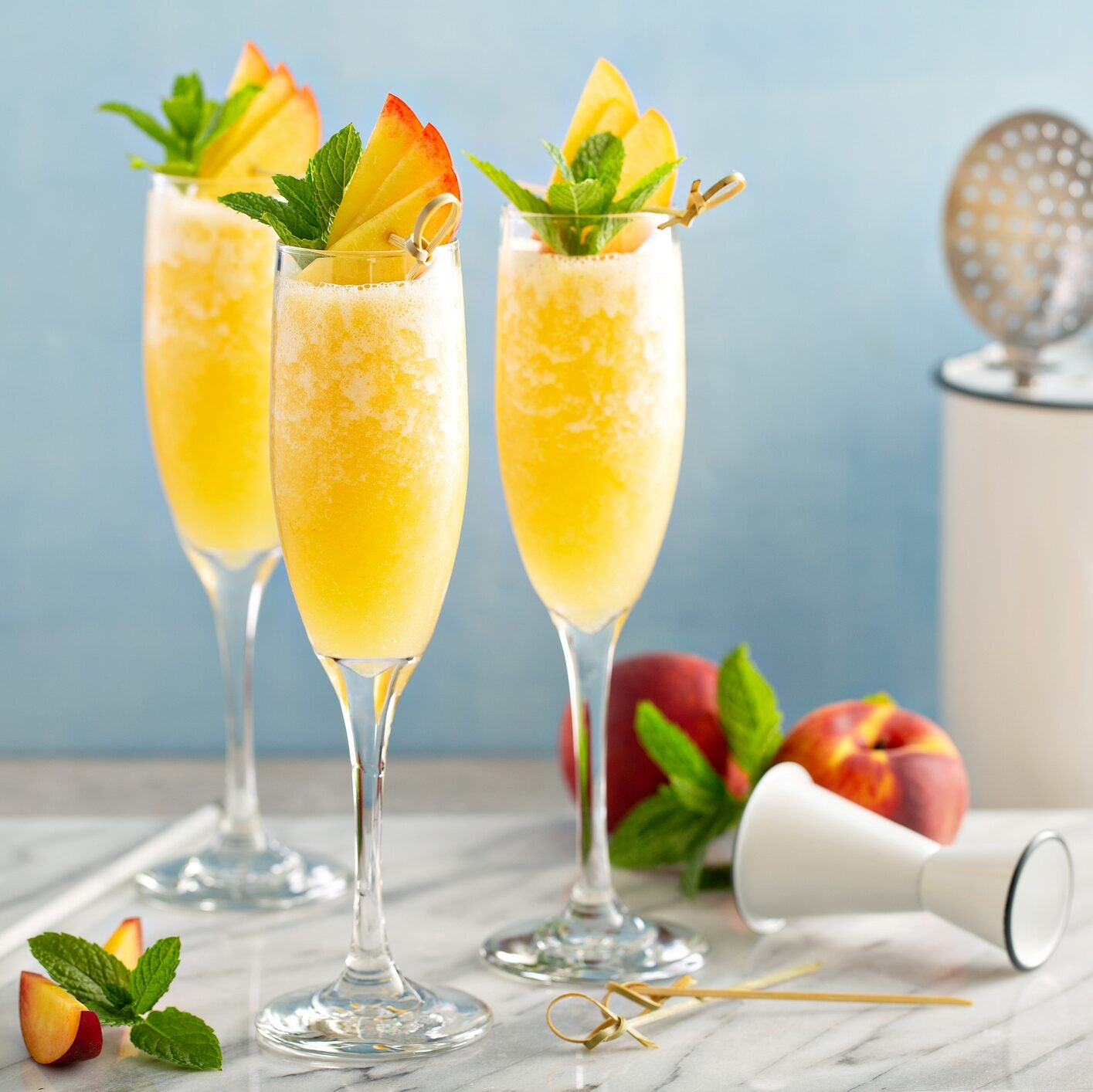 Three glasses of mimosa topped with peach slices, with a jigger, shaker, strainer and peaches on a marble countertop. 