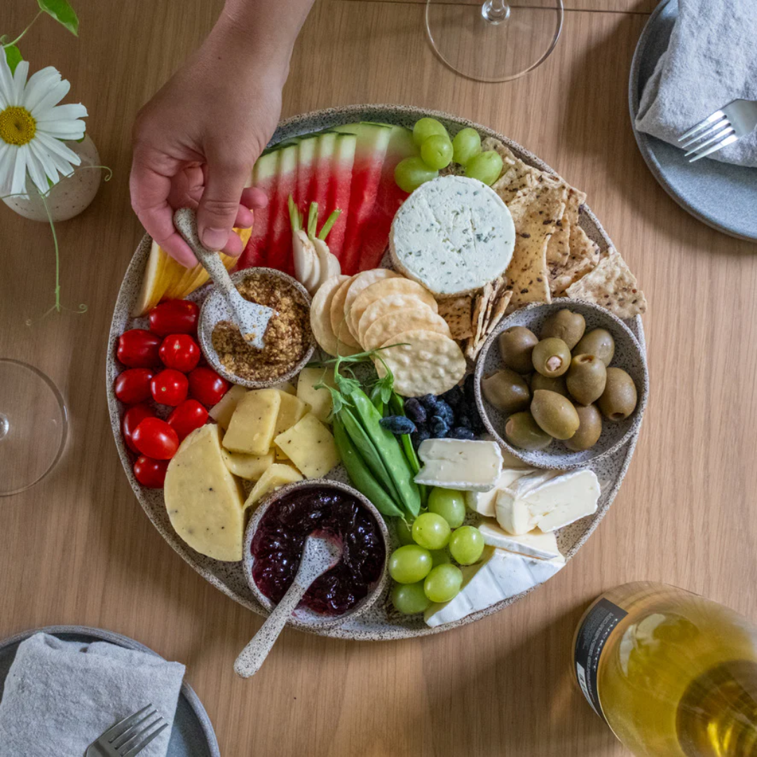 A hand holding a mini spoon dipping into a small bowl of mustard in a full charcuterie platter with cheeses and fruits.