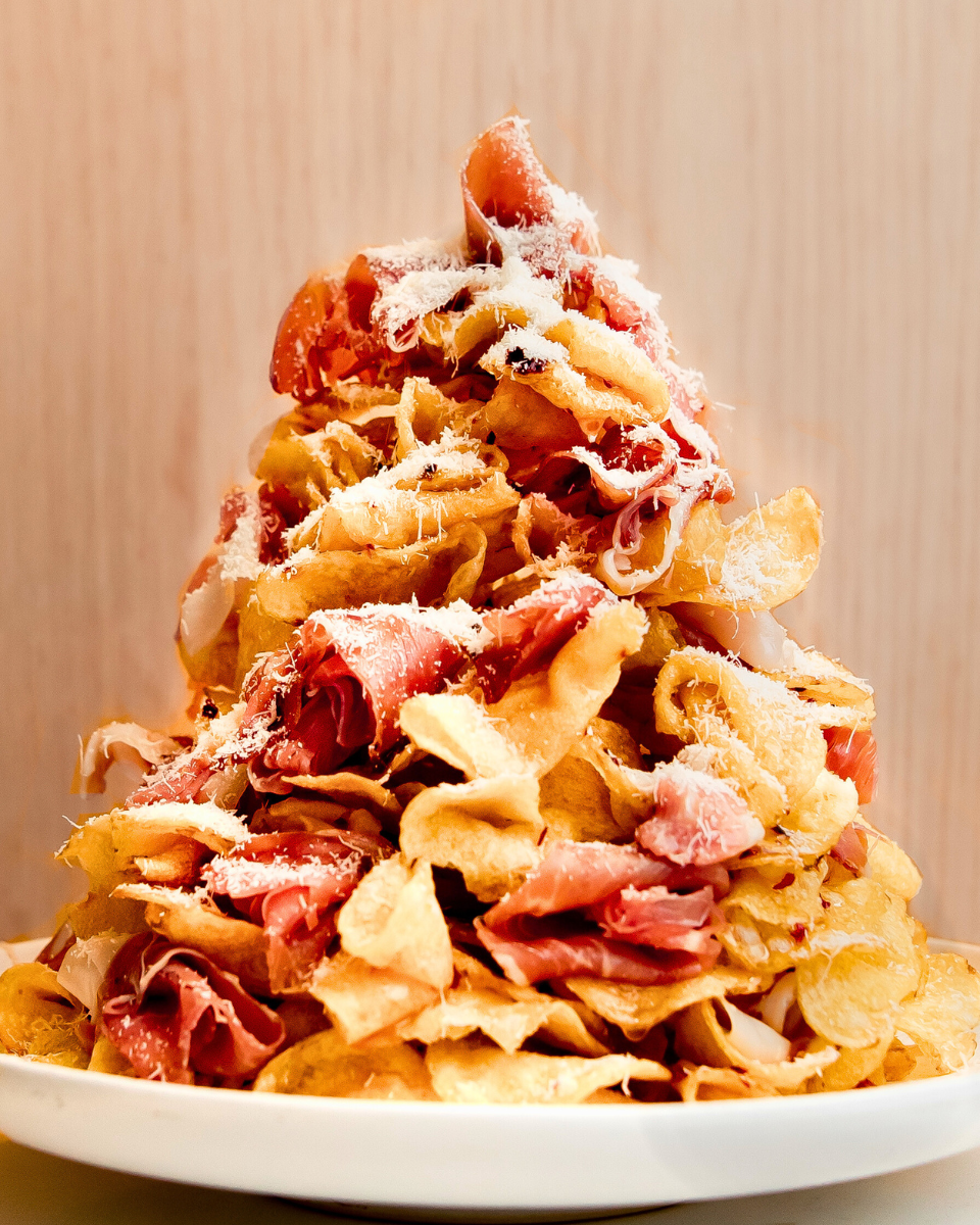 Layers of crispy kettle-cooked potato chips are stacked with torn prosciutto, drizzled with hot honey, and finished with a shower of freshly grated Parmesan.