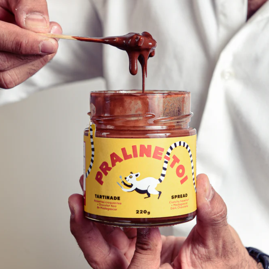 Close-up of a hand holding a spoon dripping with chocolate hazelnut spread over an open jar.