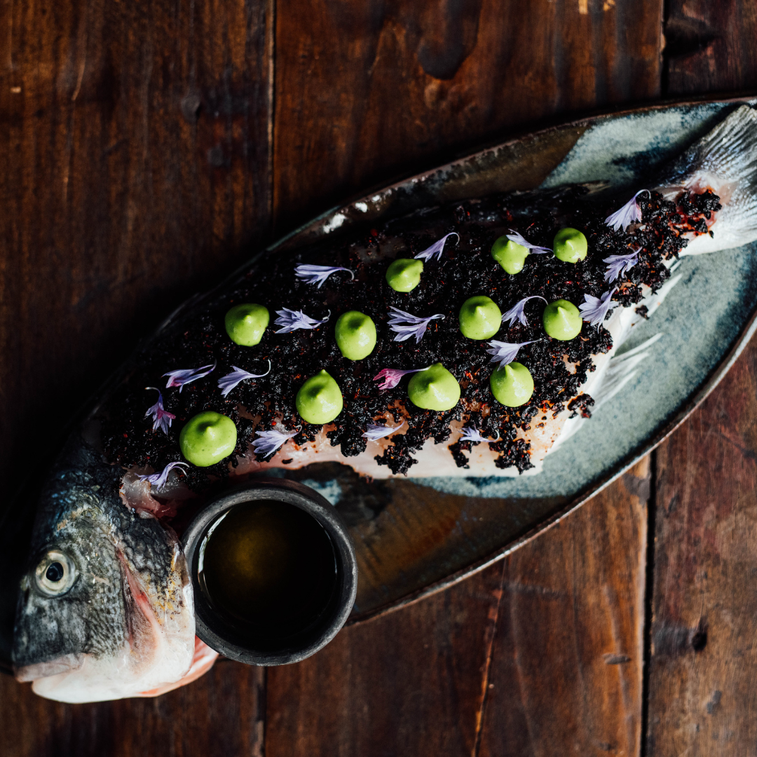 A seabream on a brown plate, with a side of sauce and topped with green dots of chile ancho sauce and hibiscus. 