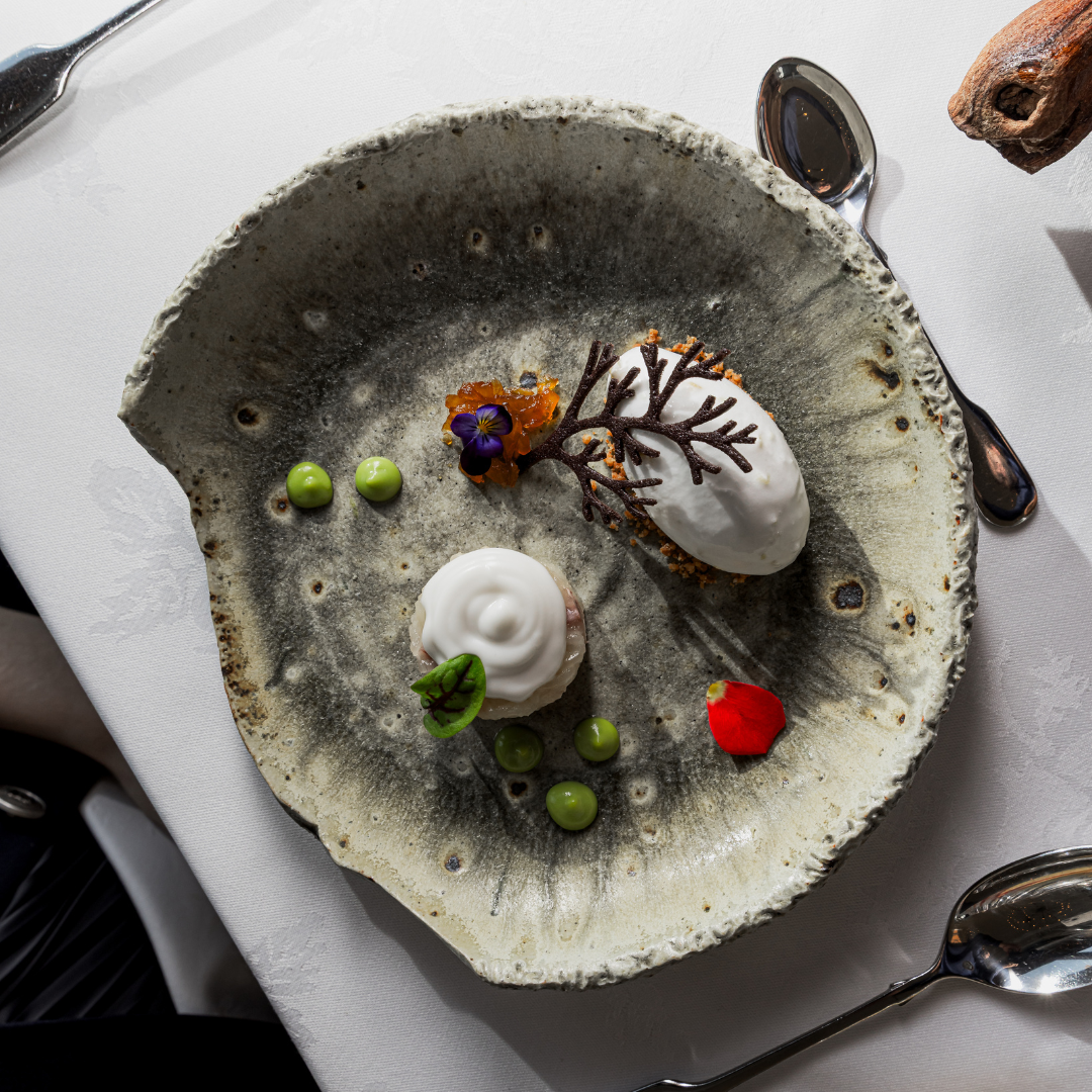A gray plate topped with a quenelle of ice cream, decorative flowers and a chocolate twig. 