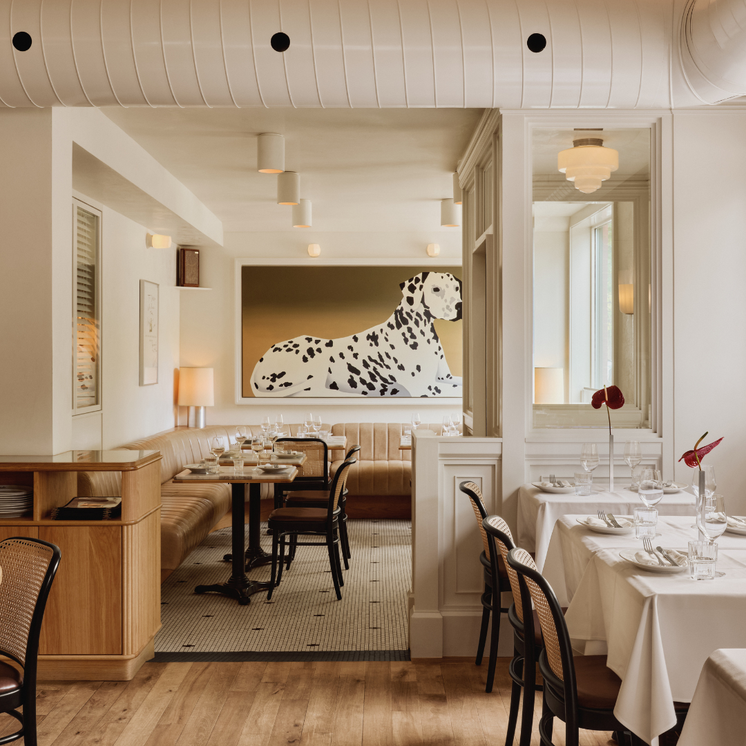 Restaurant interior with white walls and white tablecloths on tables, and a large painting of a dalmatian on the back wall. 