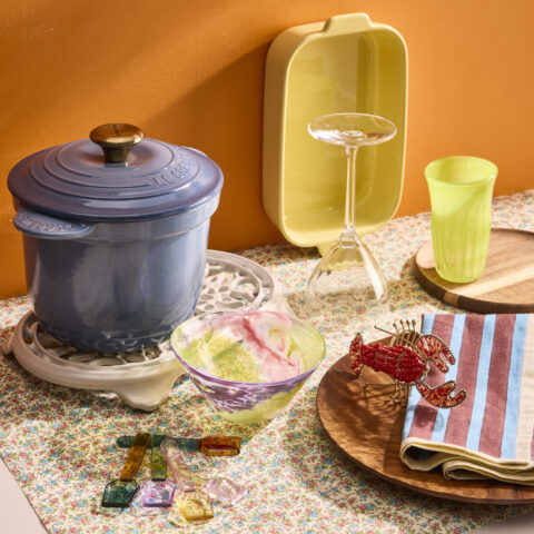 A blue ceramic pot, yellow ceramic tray, glass spoons, lobster napkin ring, glass cups and bowls on a table with orange wall background.