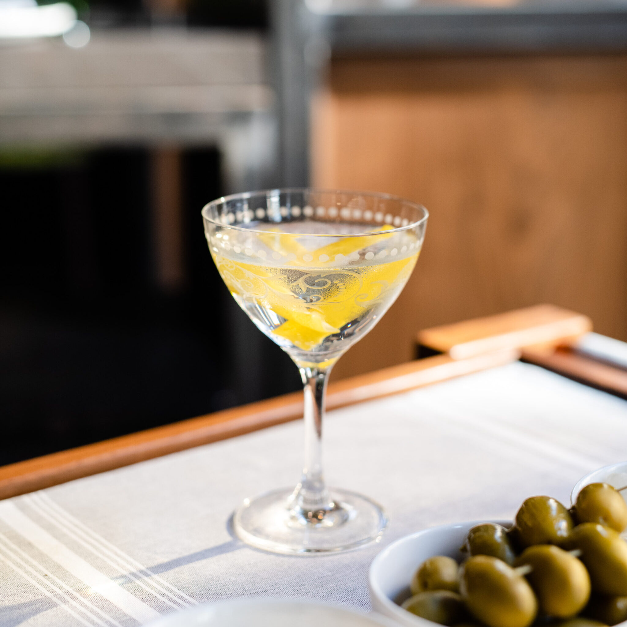 A martini in a clear glass with lemon peel next to a dish of skewered green olives.