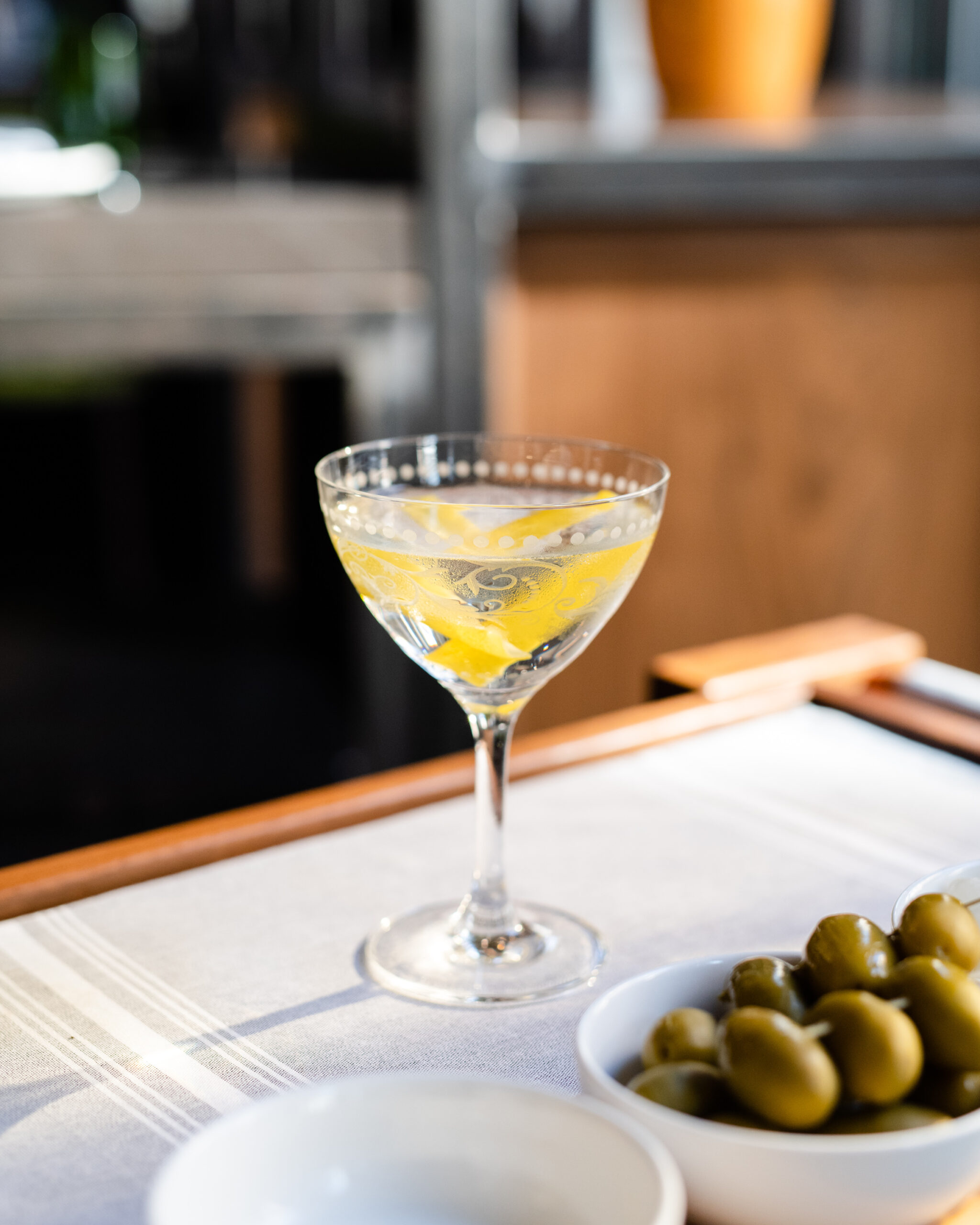 A martini in a clear glass with lemon peel next to a dish of skewered green olives.