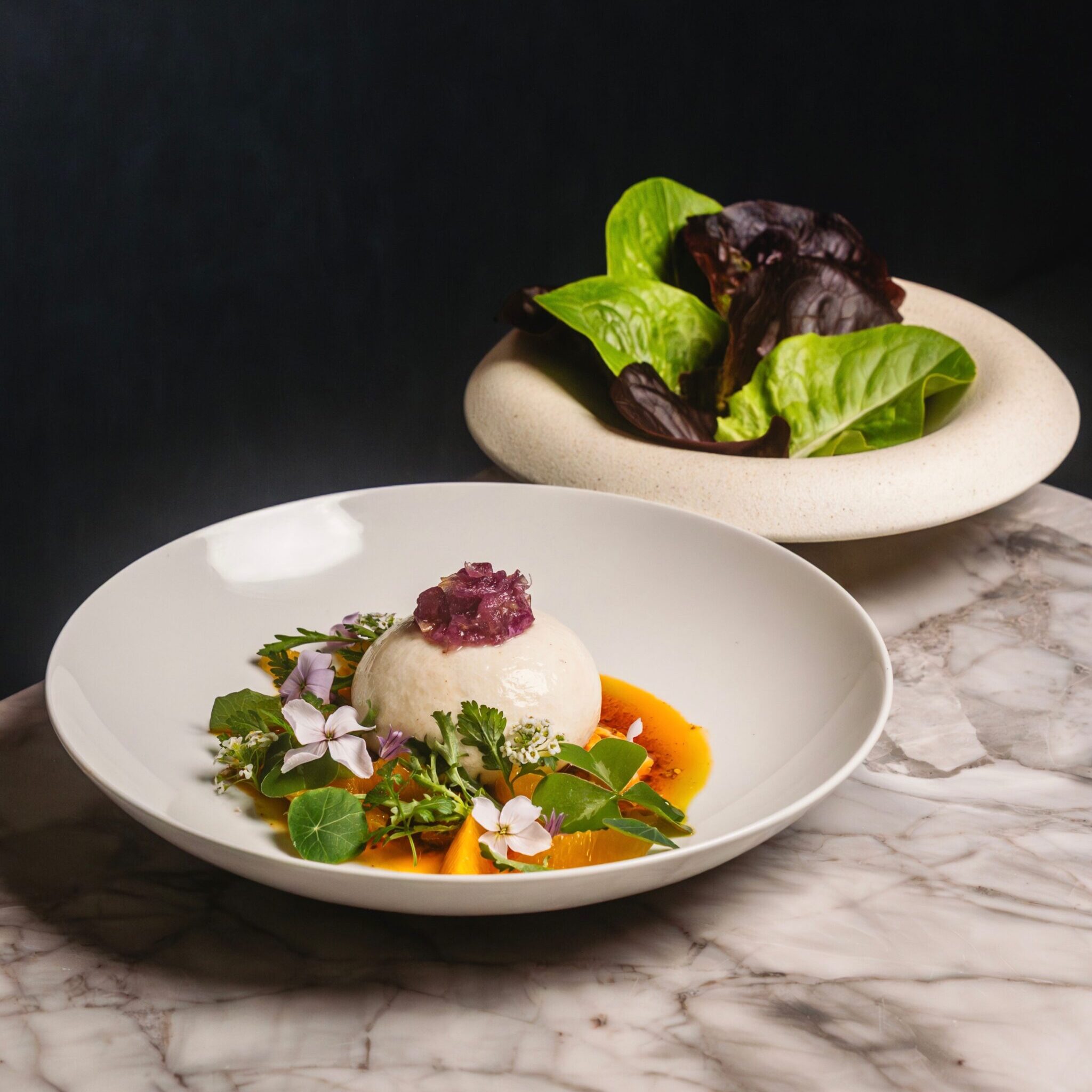 A bowl of burrata beside a bowl of lettuce on a marble countertop.