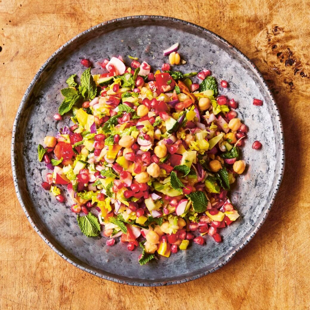 Colorful chopped salad with chickpeas, pomegranate seeds, cucumber, red onion, peppers and fresh mint served in a gray bowl on a wooden surface.
