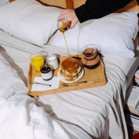A hand pouring syrup onto a stack of pancakes on a tray with orange juice and coffee on the bed.