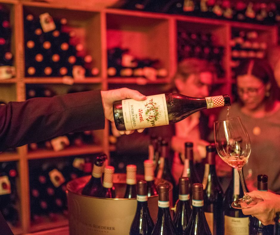 Wine being poured into a glass in a dimly lit wine cellar with bottles in the background.