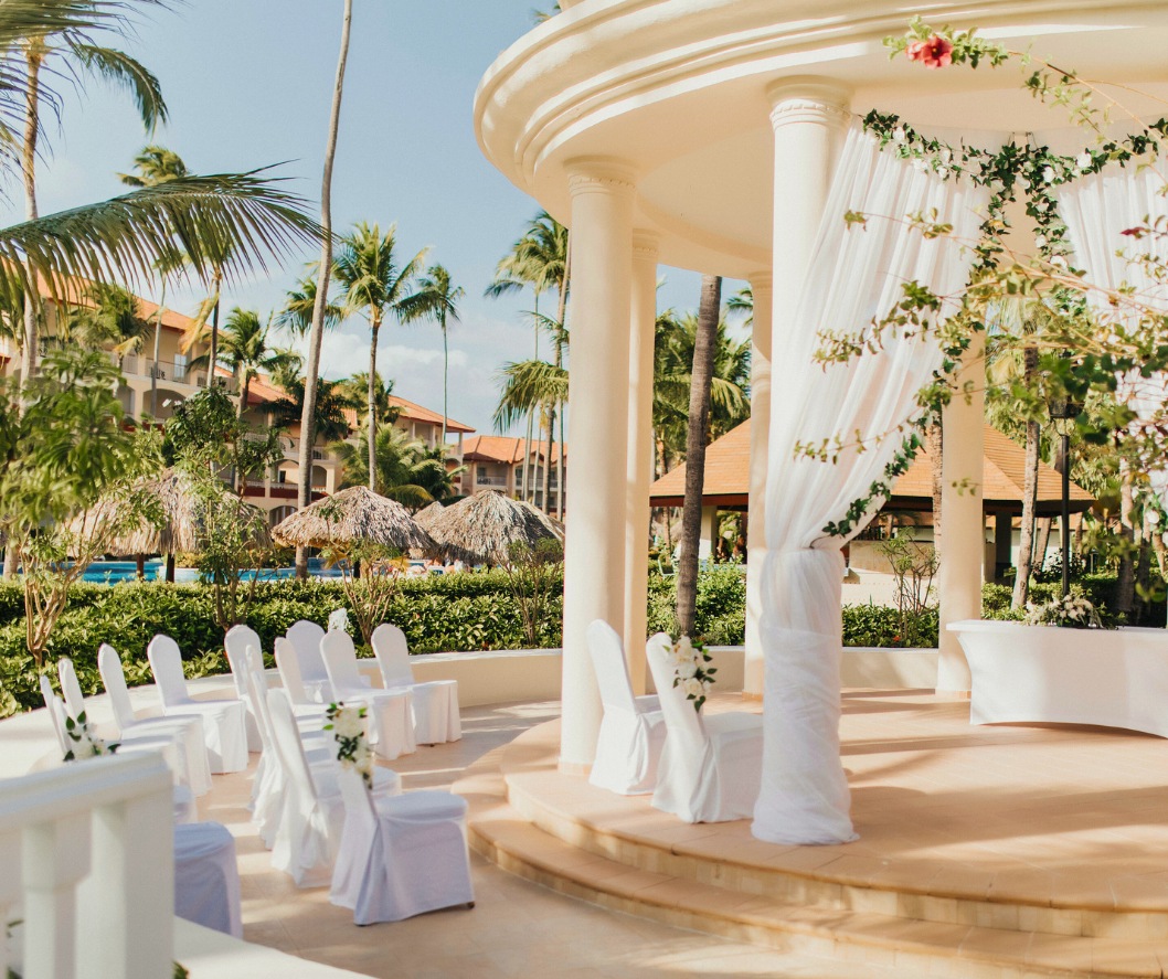 Outdoor wedding gazebo decorated with white drapery and greenery, with guest seating arranged beneath palm trees