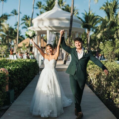 Newlywed couple walking down a garden path after an outdoor destination wedding ceremony with palm trees and a white gazebo in the background.